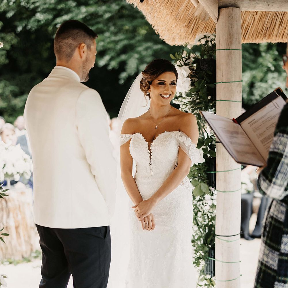 Bride in floral lace wedding dress standing at the outdoor ceremony area with the groom in a white tuxedo 