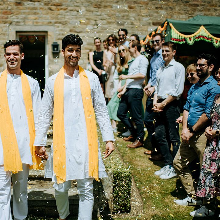 Grooms walk through petal confetti whilst holding hands and wearing matching outfits with yellow fabric draped across their shoulders