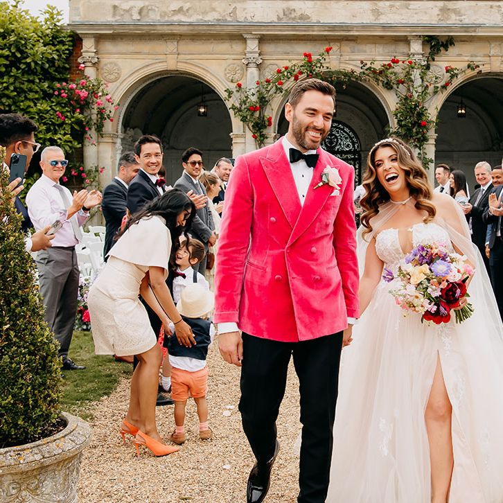 Groom in pink velvet suit jacket walking with the bride in a strapless lace wedding dress with sheer bridal cape 