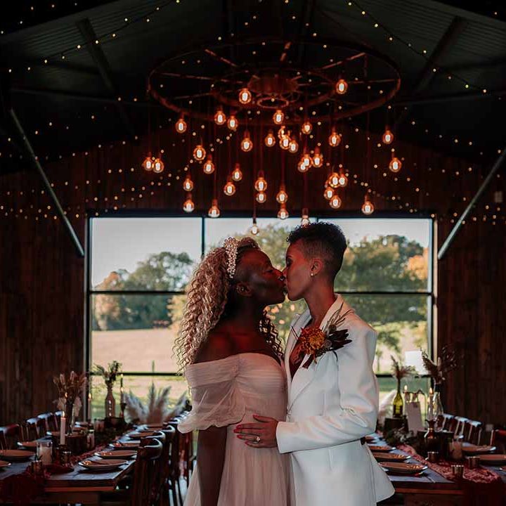 Bride in off the shoulder gold star flecked embroidered dress and bride in white silk suit with autumnal dried floral boutonniere standing in reception room of Silchester Farm wedding venue with fairy lights and festoon chandelier lighting 