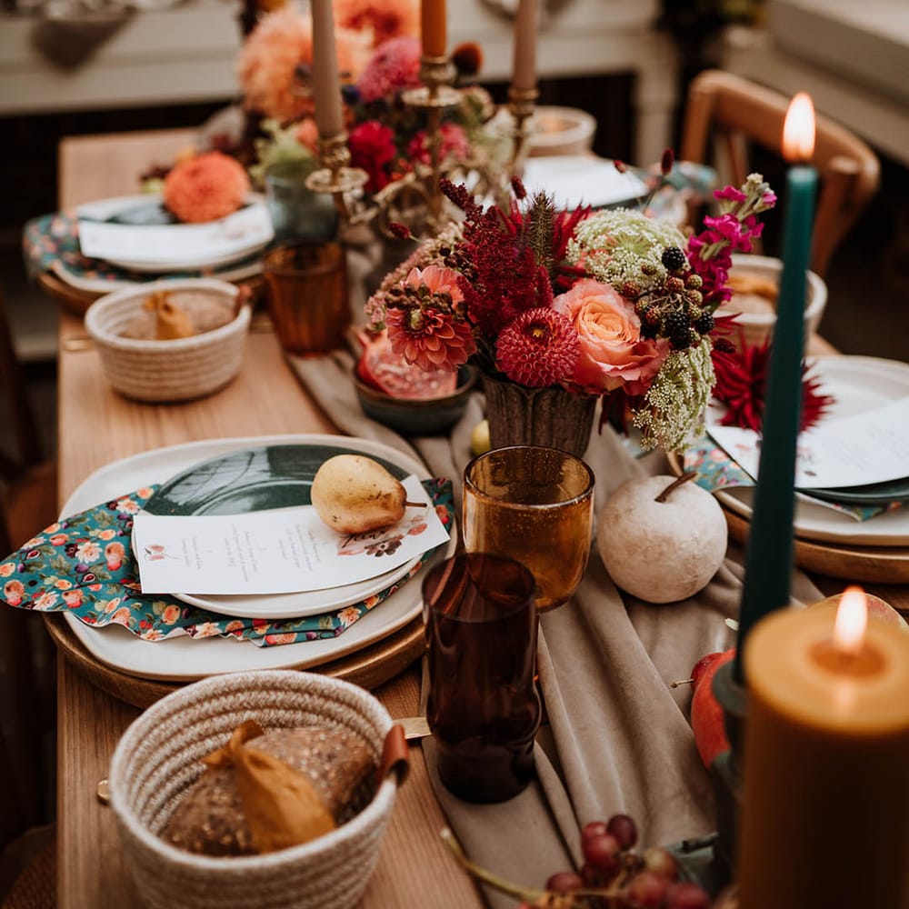 Red, brown and dark green tapered candles in gold candlesticks, tall orange pillar candles, autumnal dried flowers, seasonal fruit and vegetables, and hand painted silk napkins 