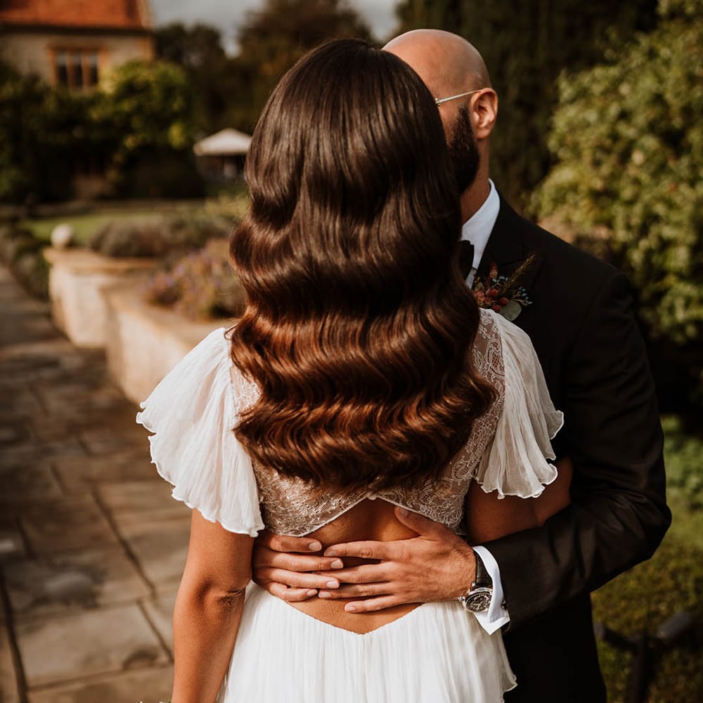 Bride wearing lace open back wedding dress with butterfly sleeves holding autumnal dried flower bouquet and groom in classic black tuxedo embracing her