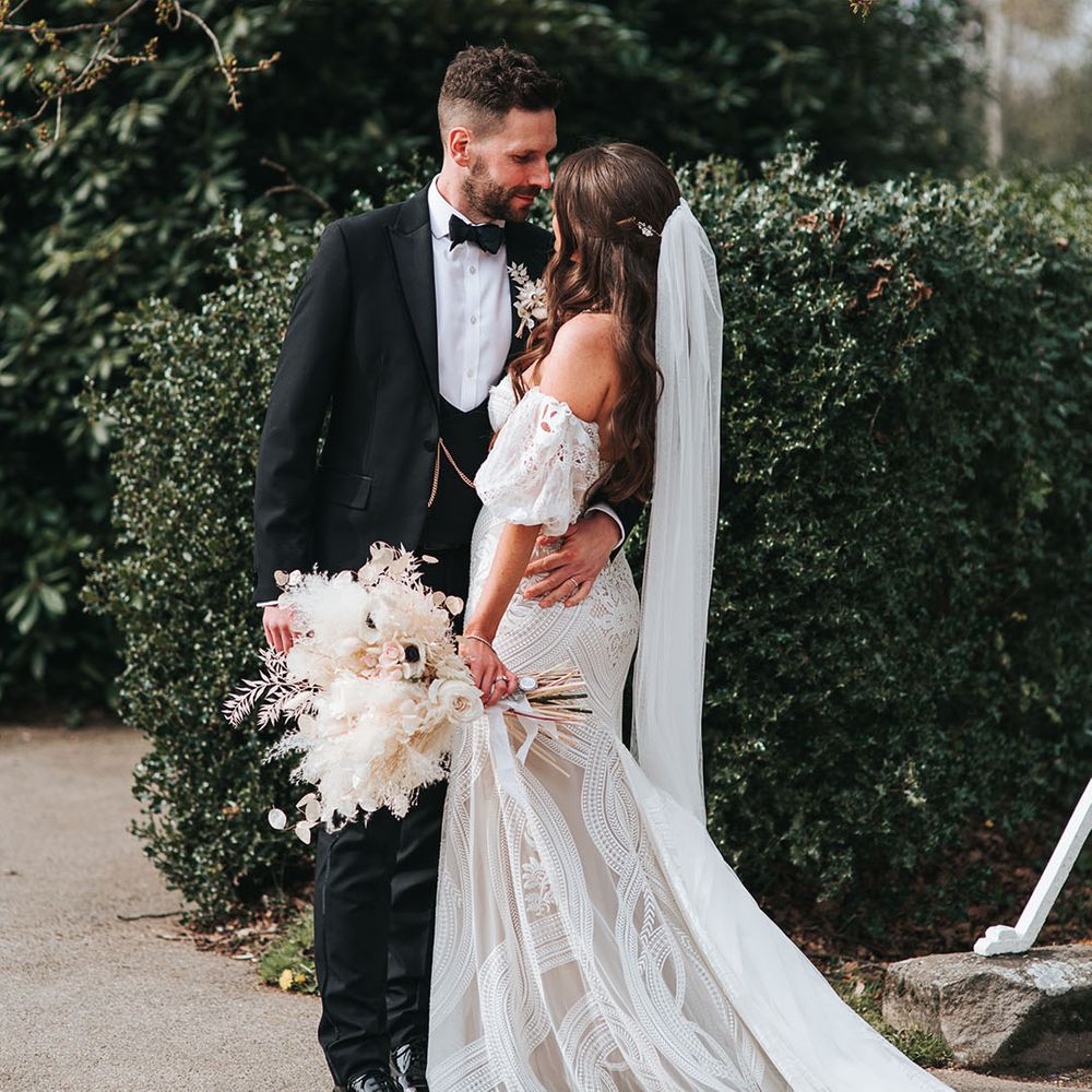 Bride with waved brown hair in a half uo half down style with veil in a boho strapless gown gazes into the groom's eyes who wears a black tux 