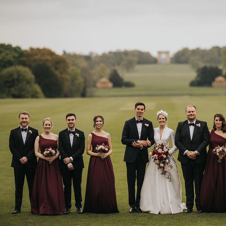 Bridesmaids in one shoulder burgundy dresses standing with the groomsmen in black tie with the bride and groom for traditional country house wedding 