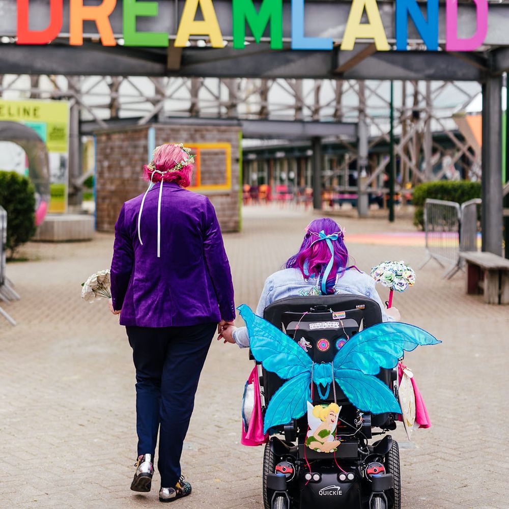Bride in wheelchair with glittery blue fairy wings and bride in purple velvet blazer and black suit trousers 