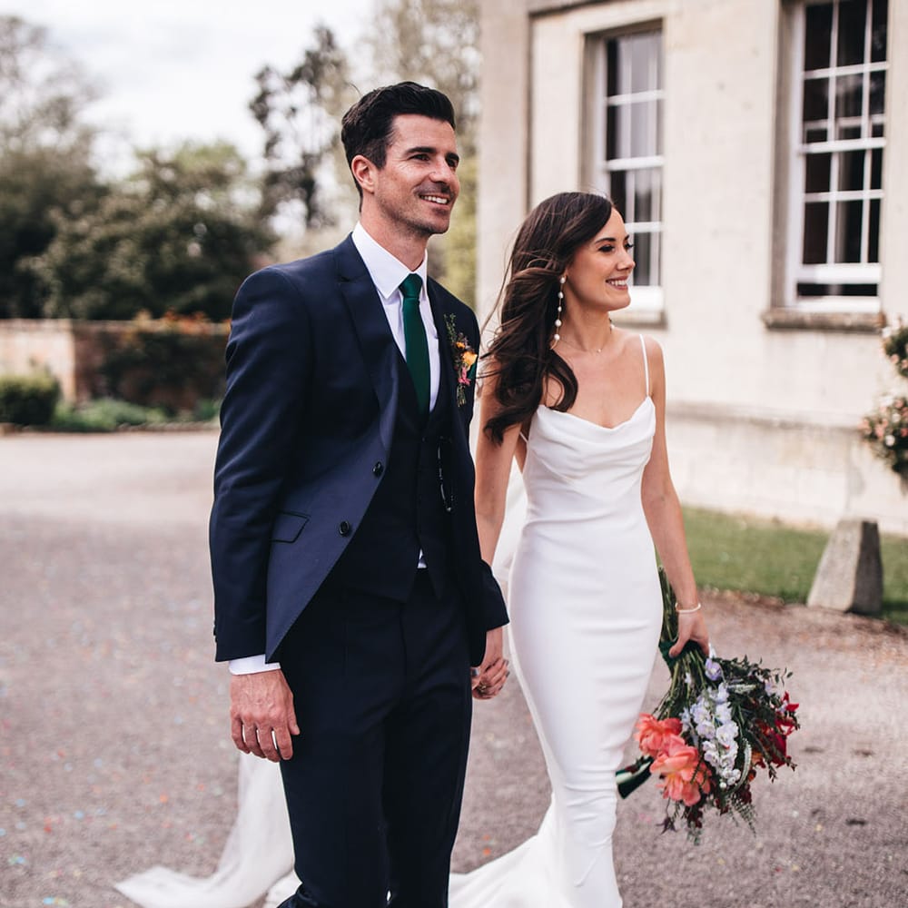 Bride with pearl earrings in Kyha wedding dress with a cowl neck and open back walking with the groom in a three piece navy suit 