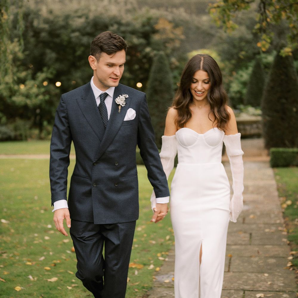 Groom in double breasted suit jacket walking hand in hand with the bride in a fitted wedding dress with detachable sleeves with pearls 