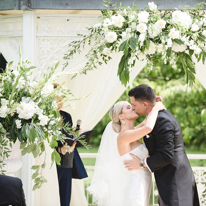 Pretty white and green floral and foliage altar decoration for outdoor wedding ceremony with the bride and groom sharing their first kiss 