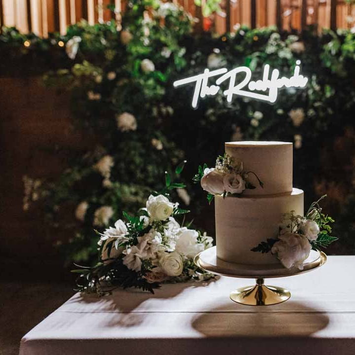 Two tiered buttercream wedding cake with garden rose and foliage decorations on golden cake stand in front of foliage wall and large neon wedding sign