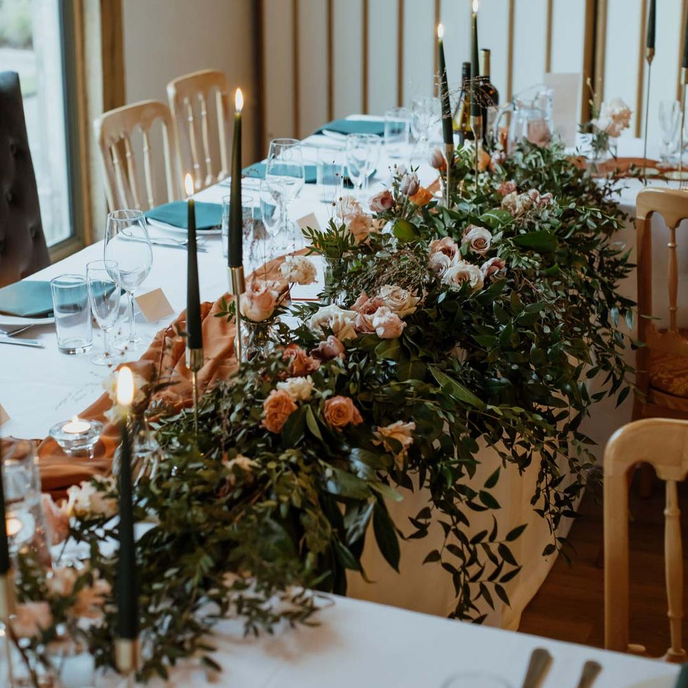 Classic wedding tablescape with white tablecloth, burnt orange wedding tablerunner, forest green wedding napkins, forest green tapered candles and bottles of wine with large light pink and orange garden rose and foliage table centrepiece