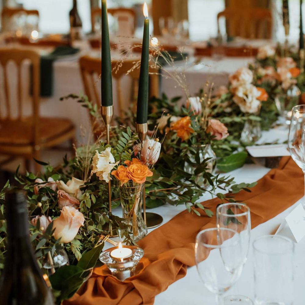 Burnt orange wedding tablerunner, forest green wedding napkins, forest green tapered candles and bottles of wine with suspended foliage and fairy lights decor at wedding