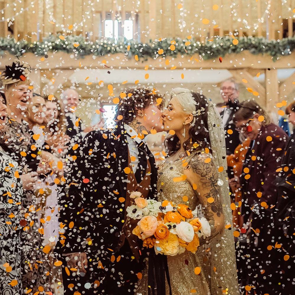 Bride in dark velvet bridal jacket with rust waistcoat and pocket square, black suit trousers and gold pocket watch kissing bride in sleeveless deep v neck sparkly gold wedding dress with sheer and gold polka dot wedding veil holding orange garden rose, white poppy, light pink carnation and foliage bridal bouquet wrapped in sparkly gold ribbon during colourful confetti exit at Kingscote Barn Tetbury wedding venue with fairy lights and festoon lighting