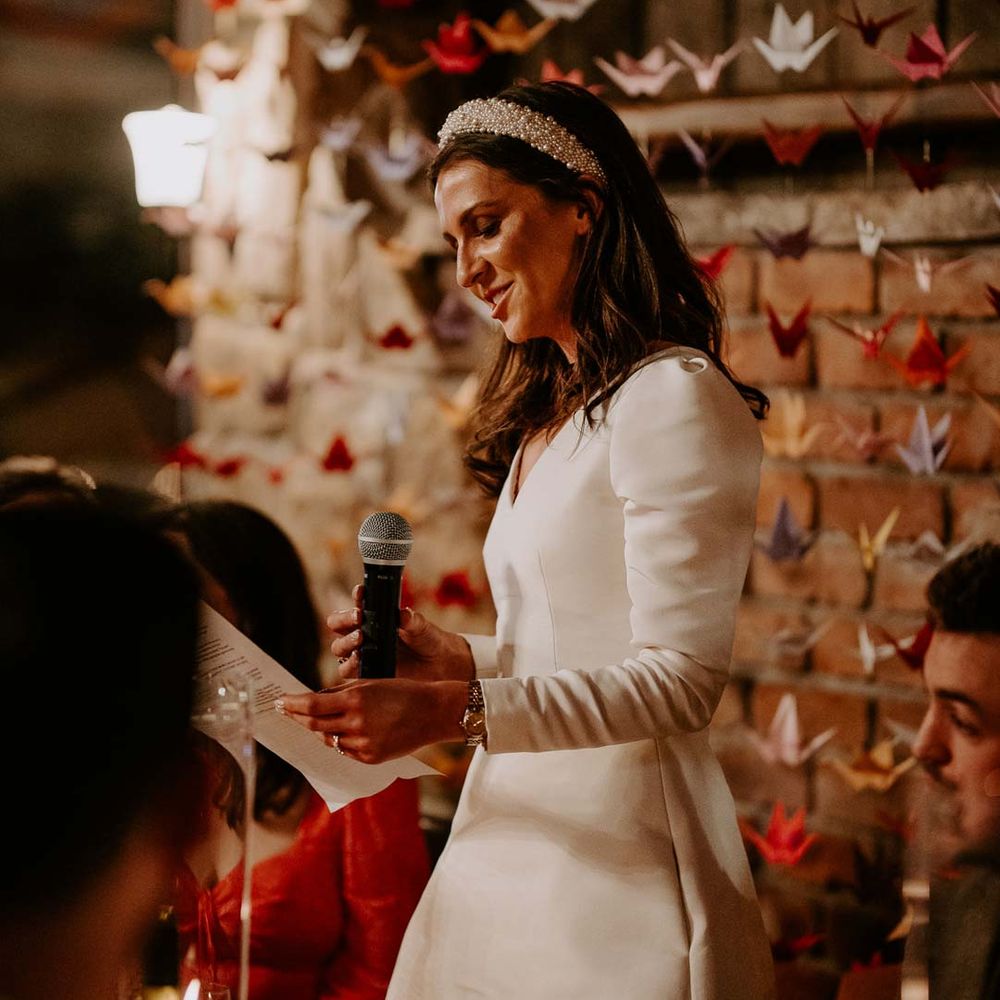 Bride in long sleeve short wedding dress and pearl bridal headband doing bridal speech standing in front of origami paper cranes DIY wedding decor at Willow Marsh Farm Loughborough 