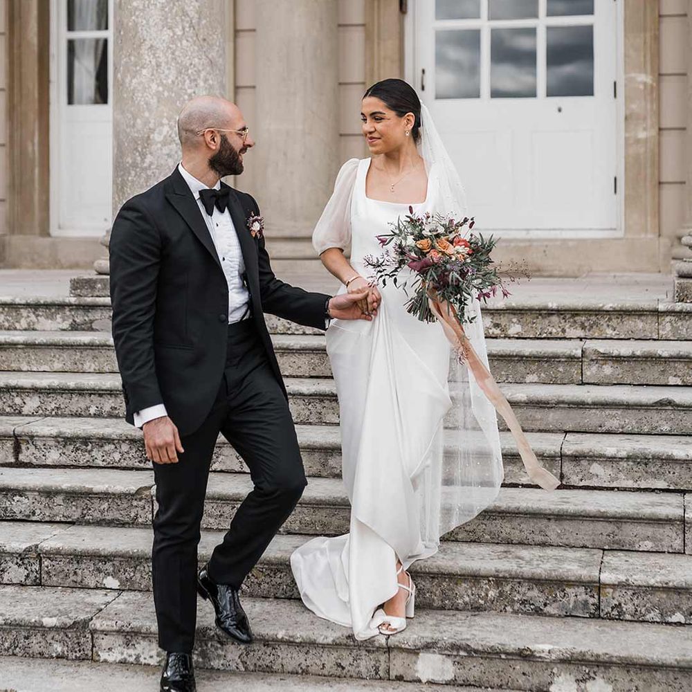 Groom in classic black tuxedo with bowtie and wildflower boutonniere walking hand in hand with bride in short sheer puff sleeve wedding dress with square neck and wrist loop with sheer pearl embellished bridal veil holding dried wildflower bouquet at Buxted Park