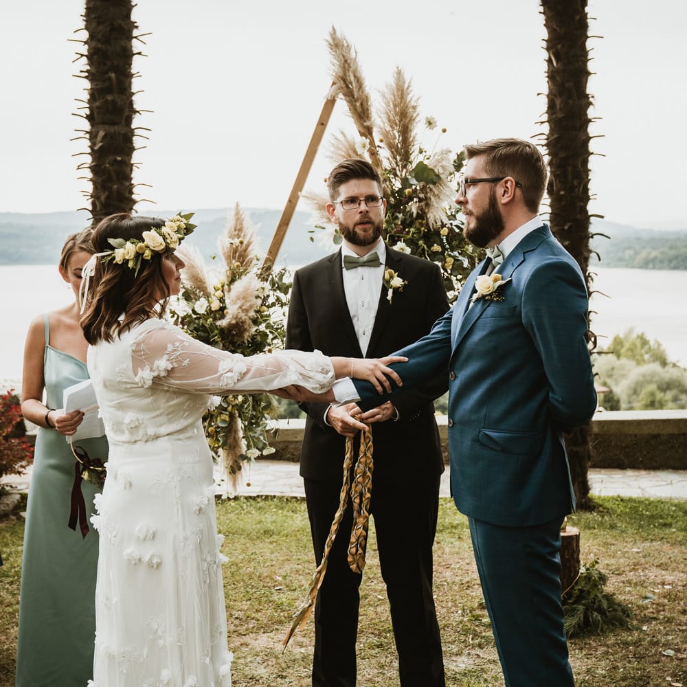 Handfasting wedding ceremony with boho bride in a flower crown and white wedding dress with flowers and the groom in a blue suit and bow tie