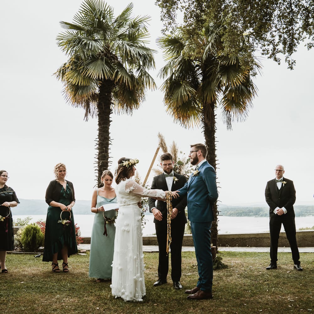 Bride in a white wedding dress with flowers and groom in a blue suit taking part in their hand fasting ceremony at Cella Grande, Lake Viverone, Italy