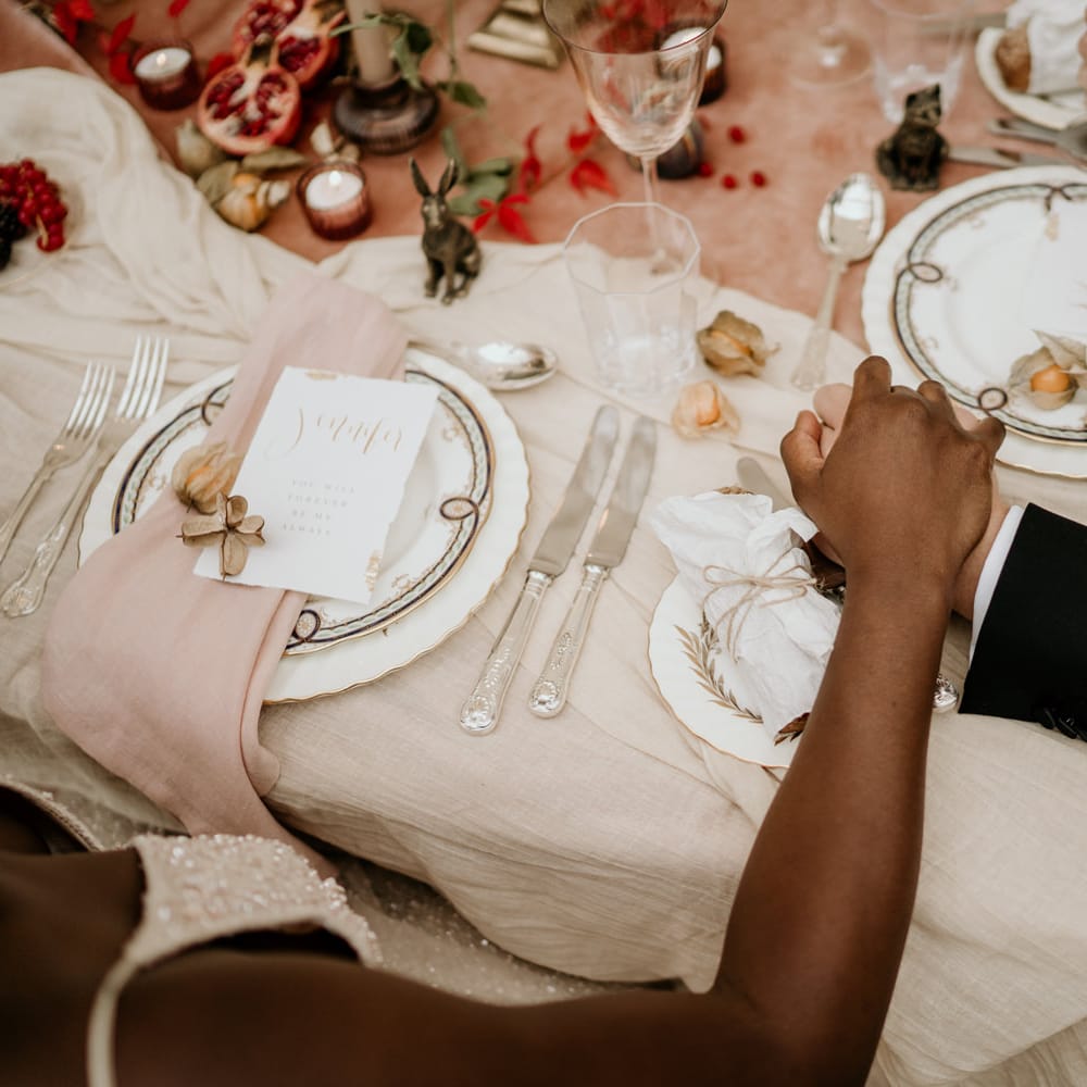 Bride and groom holding hands at their sweetheart table with ornate tableware, silver cutlery and natural linens 