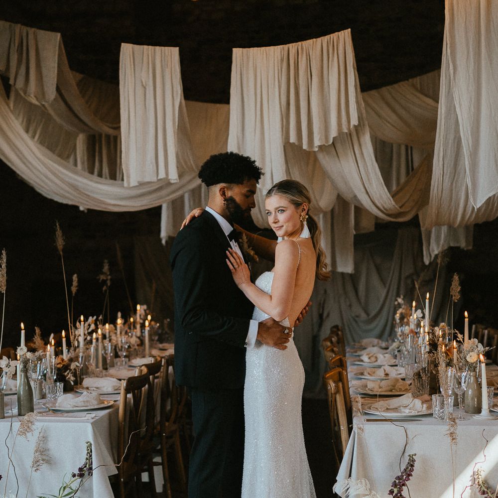 Groom in a tuxedo and bride in a sequin wedding dress standing in their Pentney Abbey wedding reception with ceiling drapes 
