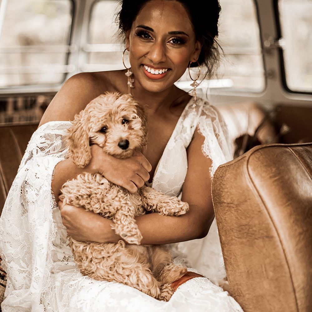 Indian bride in a lace wedding dress and hoop earrings holding a tan cockapoo puppy 