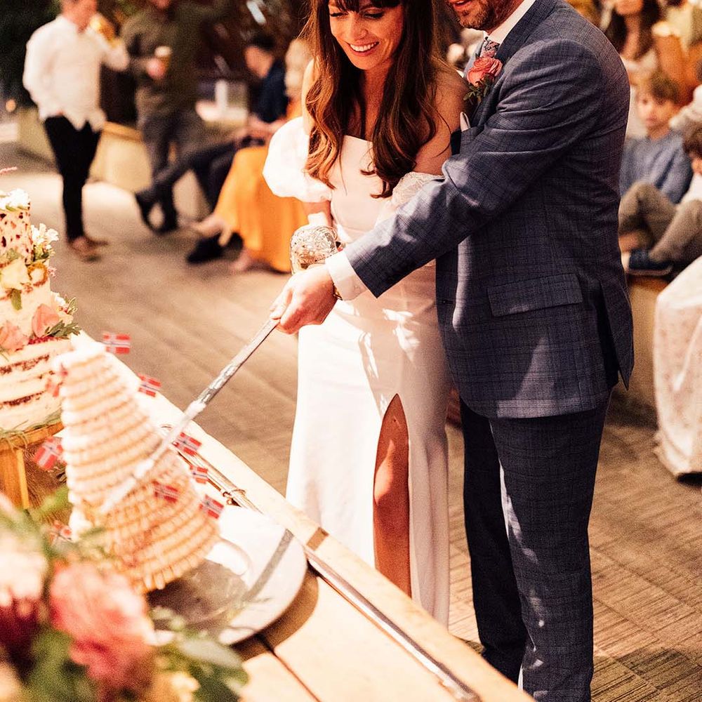 Bride in a strapless wedding dress and groom in a blue check suit cut cake with a long sword on their wedding day 