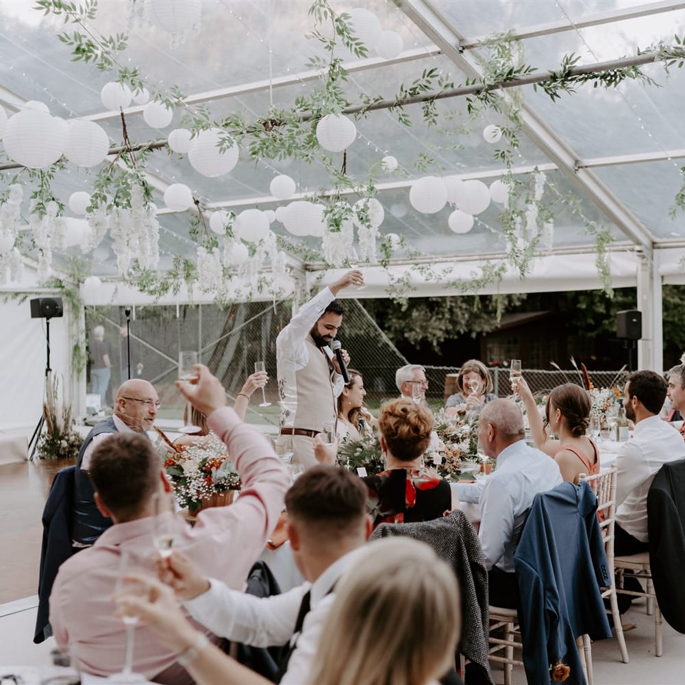 Groom in beige suit and white shirt holds dunk in the air as wedding guests raise their glasses during marquee wedding reception