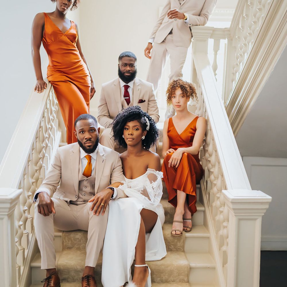 Bride with naturally curly hair in a strapless wedding dress linking arms with her groom in a beige suit on the staircase at Modern Hall with their groomsmen and bridesmaids behind them 
