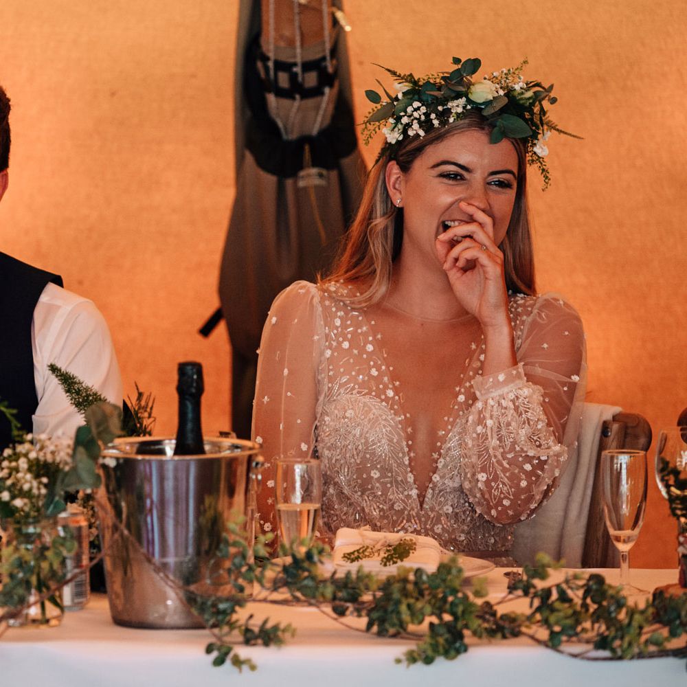 Bride in v neck embroidered Julie Vino wedding dress and boho flower crown sits laughing with groom in red tie and waistcoat as they sit at top table for tipi wedding reception