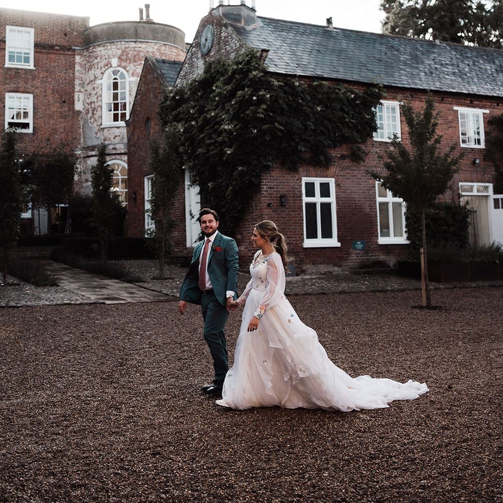 Groom in blue suit and pink tie walks holding hands with bride in bespoke Victoria Sanders wedding dress with train in the grounds of Iscoyd Park