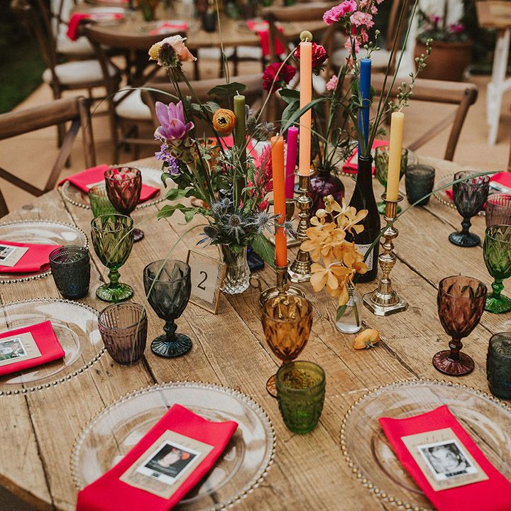 Round wedding reception table with clear glass charger plates, pink napkins, polaroid name place cards and colourful candles, vases and flower stems centrepiece decor 