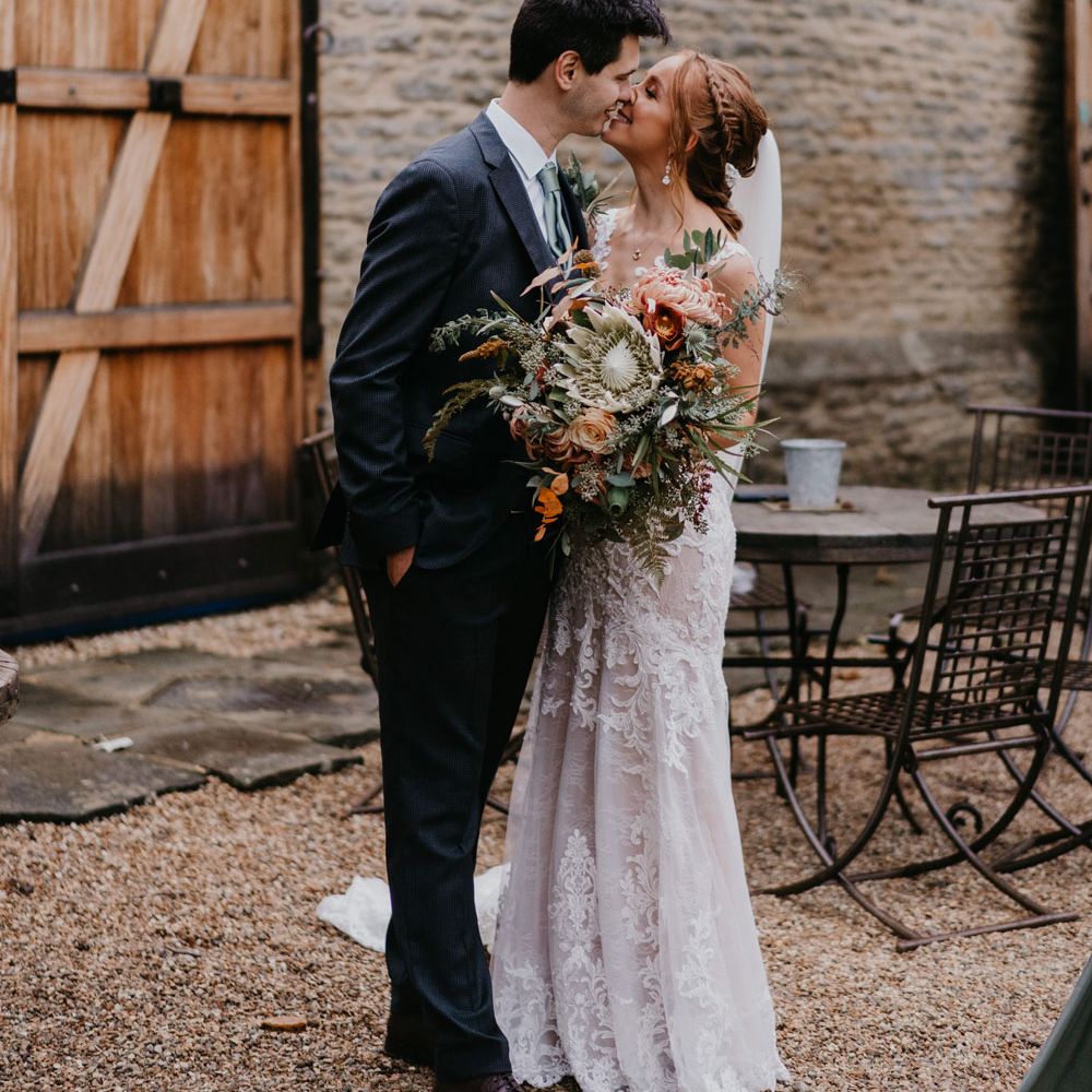 Bride in lace and tulle wedding dress with veil holding large bridal bouquet kisses groom in dark suit in barn courtyard 
