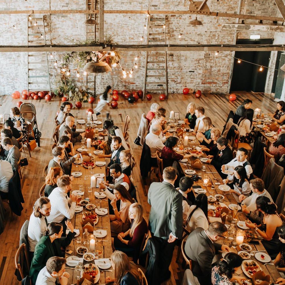 Wedding guests sit at long wooden table in industrial style wedding reception room with beams and exposed brick 