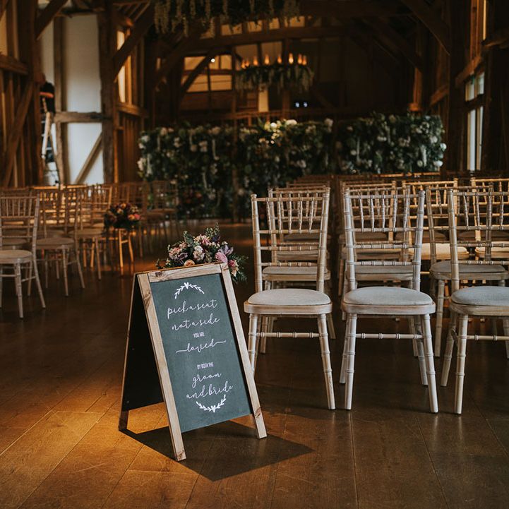 Rustic wedding venue with light wooden and cream seats with flower displays and chalk notice board reading 'Pick a seat, not a side. You are loved by both the bride and groom.'