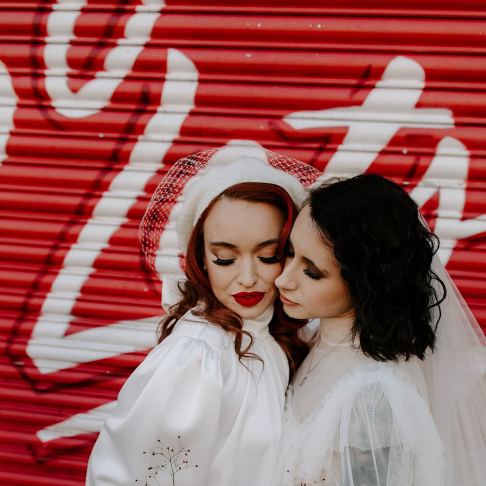 Two brides embracing in tulle and satin short wedding dresses with high necks holding a red and pink romantic wedding bouquet 