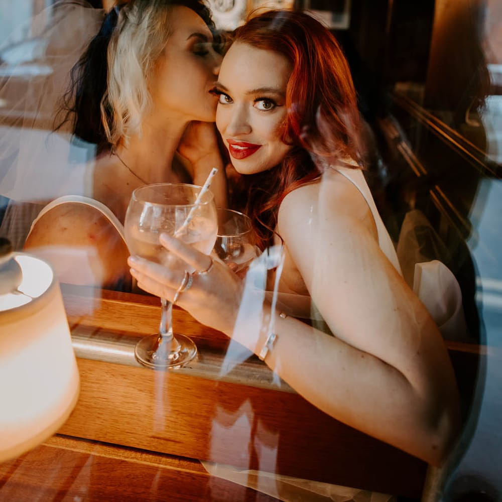 Bride with two tones hair kissing her bride with red hair as she sips gin from a balloon glass 