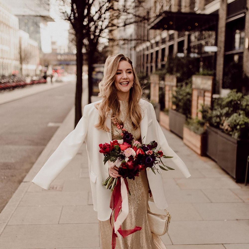 Bride in gold wedding dress and small handbag with white blazer cover up and pink, red and purple wedding bouquet 