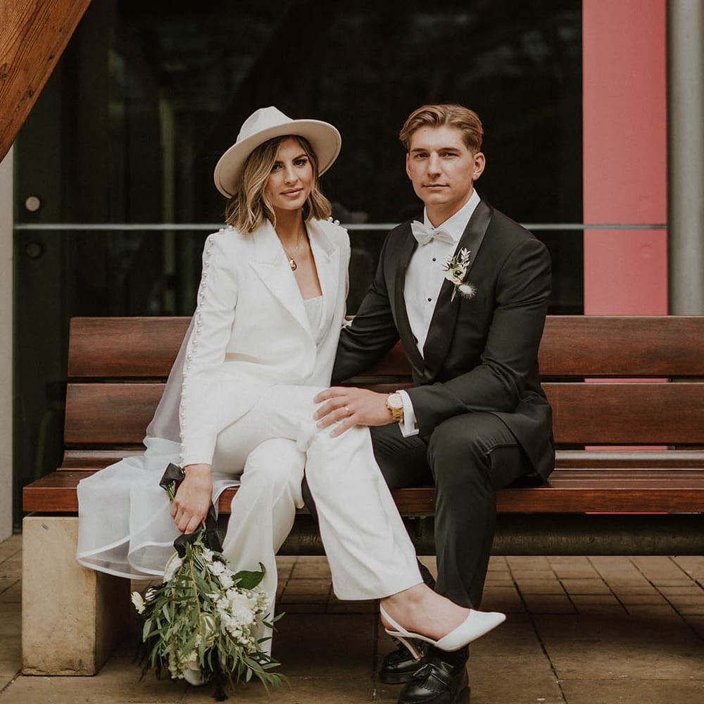 Groom sits in black tie with his hand on the bride's knee as she wears white trouser wedding suit and white slingback heels