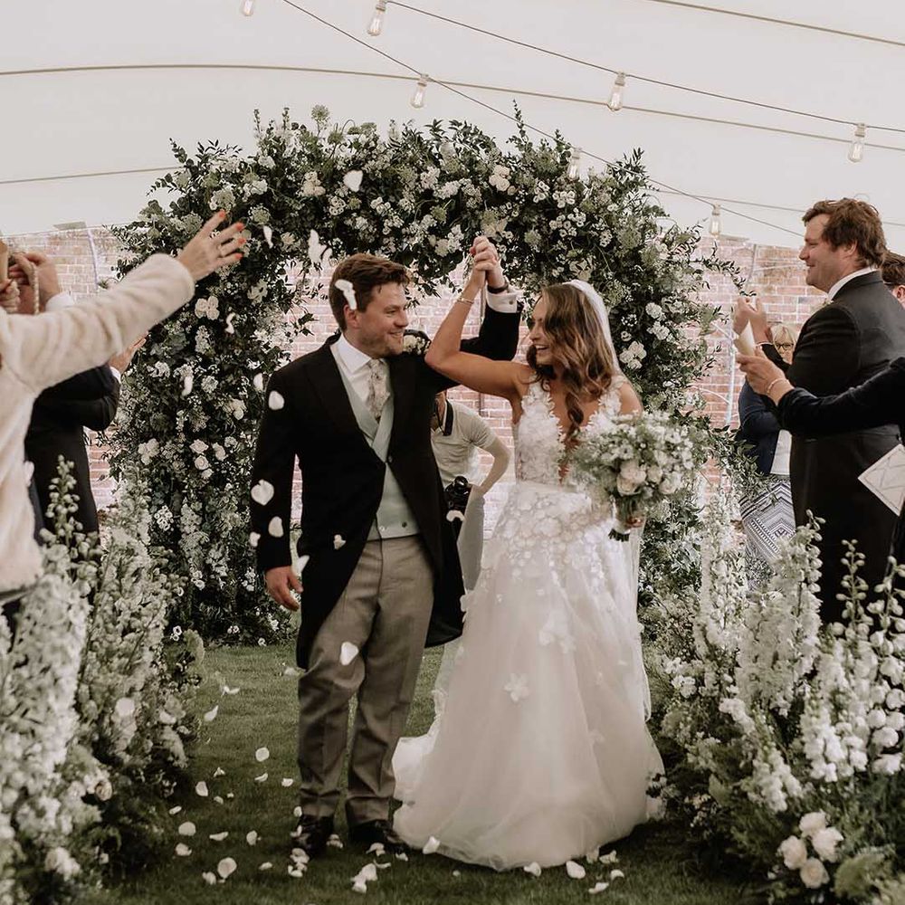Bride and groom lift their intertwined hands as they walk back down the aisle to confetti as a married couple