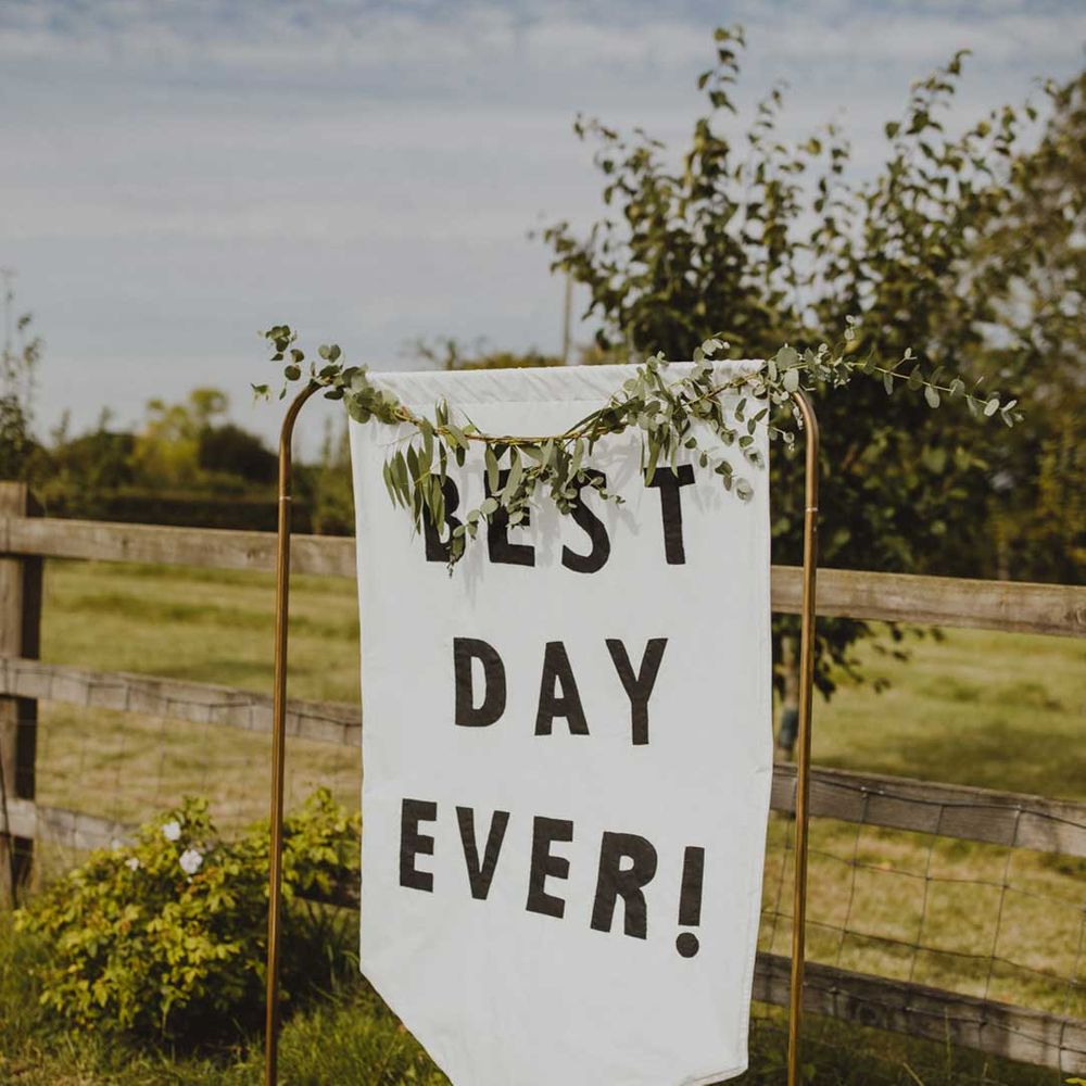 DIY white fabric wedding sign reading "Best Day Ever!" on copper sign stand with floral details around the sides