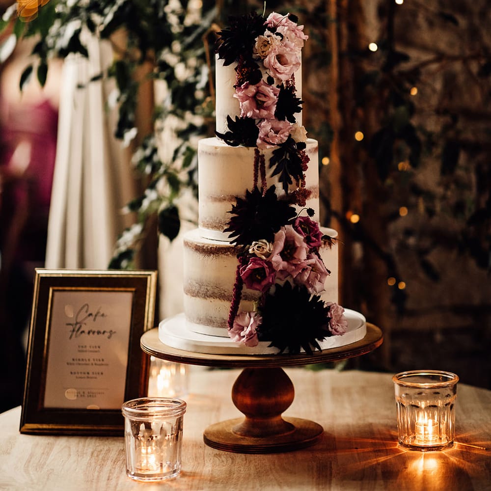 Three tier semi naked wedding cake decorated with dark flowers on a wooden cake stand for the rustic wedding 