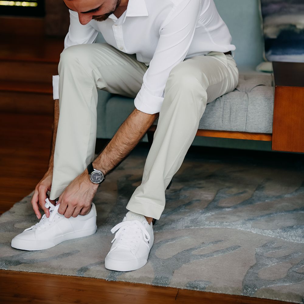 Groom in crisp white shirt, light coloured chinos, tan leather belt tying up his white trainers