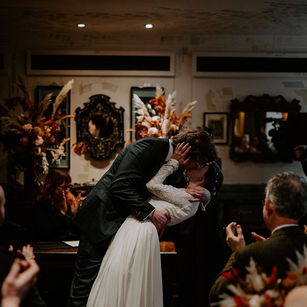 Bride in long sleeve lace vintage wedding dress and closed toe satin heels kissing groom in dark suit with orange tie at the alter of Autumnal wedding