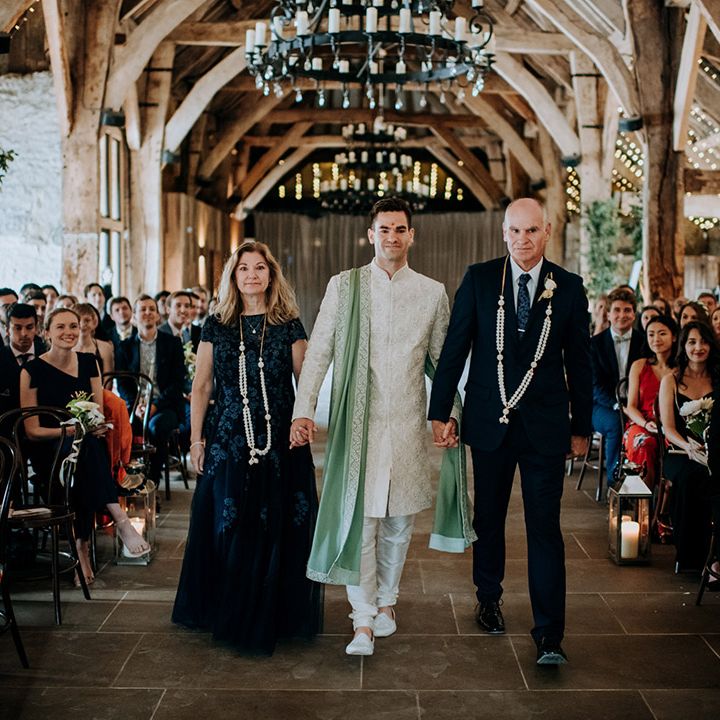 Groom in sage green sherwani walks down the aisle with his mother and father during barn wedding 