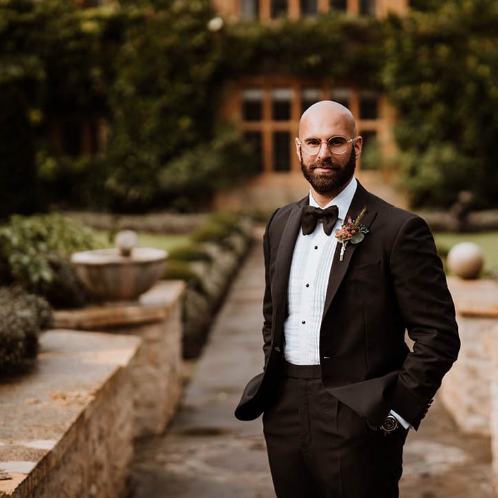 Groom in the garden of Oxfordshire wedding venue wearing classic black tuxedo, autumnal boutonniere and bowtie