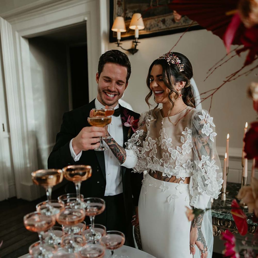 Groom in classic black tuxedo with magenta amaryllis and orchid boutonniere and bride in two piece bridal separates with puff sleeve sheer crop top with 3d appliqué flowers and full bodied silk skirt having a glass of pink Champagne next to their wedding champagne tower 