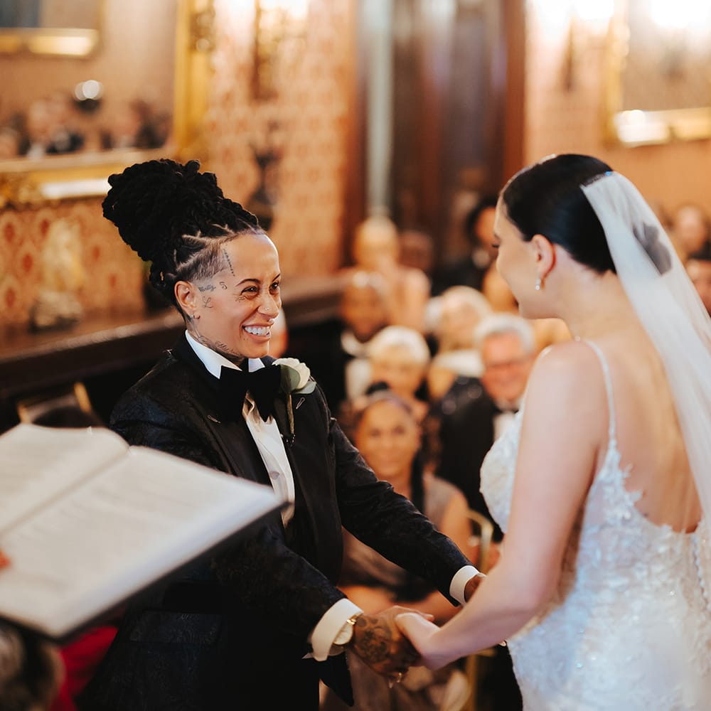 Bride in black snakeskin print tuxedo with black bowtie and white rose boutonniere holding hands with bride in lace sleeveless v neck Enzoani wedding dress with tulle finish and church length veil at Grittleton House