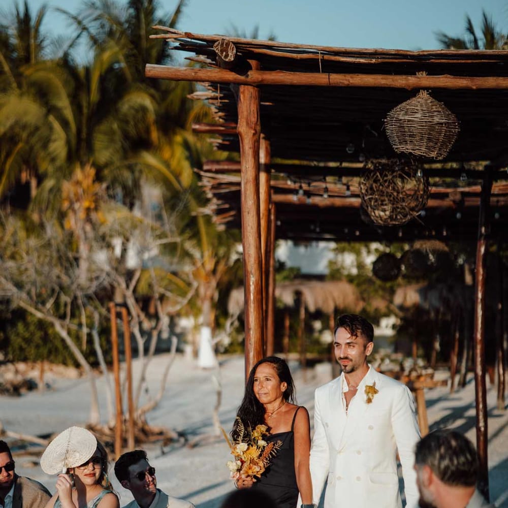 Groom walking down the aisle in white mens wedding suit, dusky orange garden rose and dried flower boutonniere, white shirt, and silver jewellery with wedding guest in black satin strappy dress 