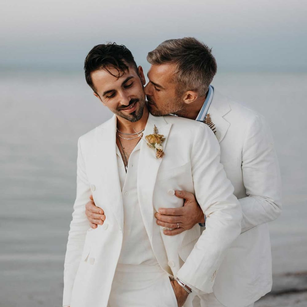 Groom in mens white wedding suit with dusky orange garden rose and dried flower boutonniere, white shirt and silver jewellery standing on the beach with Groom wearing white blazer, light blue and white striped shirt and dusky orange garden rose and dried flower boutonniere