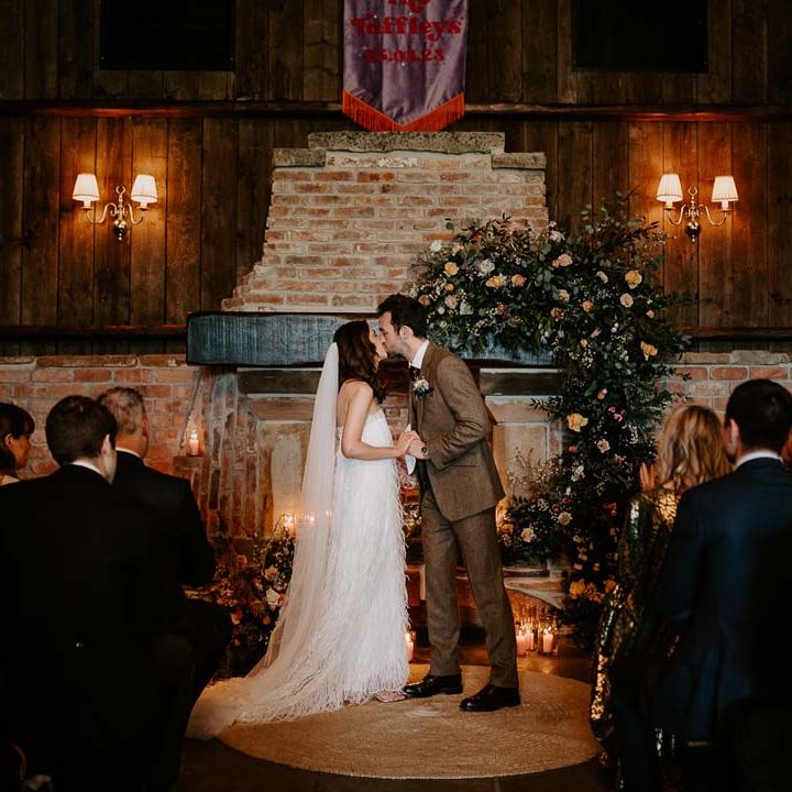 Groom in chocolate brown grooms suit kissing bride in strapless ivory fringed crepe gown and chapel length veil at the alter of Willow Marsh Farm Loughborough 