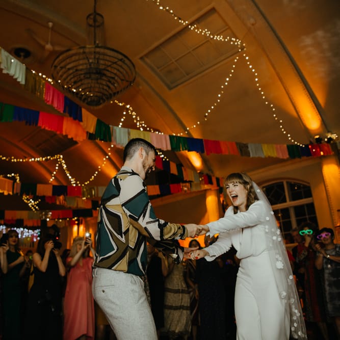 Groom in grey trousers and a grey and mustard pattern shirt dancing with his bride in a jumpsuit and appliqué veil 