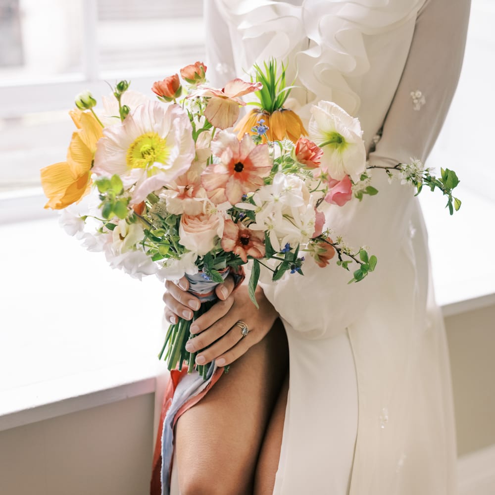 Bride holds bright & bold pastel bouquet tied with ribbon and wears pearl jewellery 